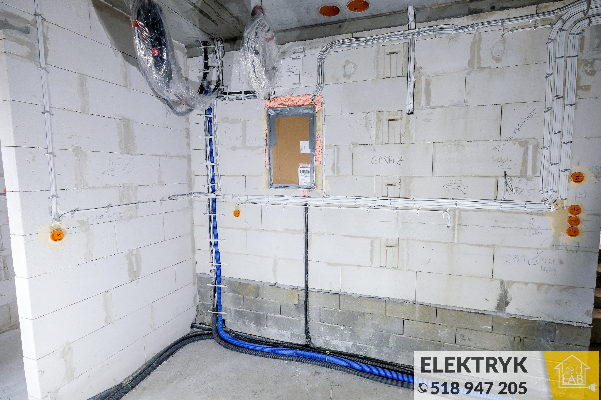 Surowy stan budynku z widocznymi, poprowadzonymi przewodami elektrycznymi w białych listwach i niebieskich rurach karbowanych, otwory pod gniazdka w ścianie z betonu komórkowego.