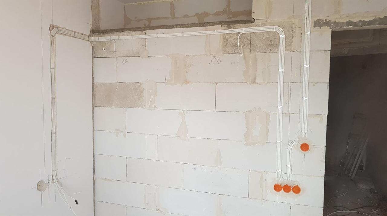 Białe przewody elektryczne prowadzone natynkowo po ścianie z białych bloczków i płyty gipsowej, pomarańczowe puszki instalacyjne w ścianie, widoczne kable wystające ze ściany.