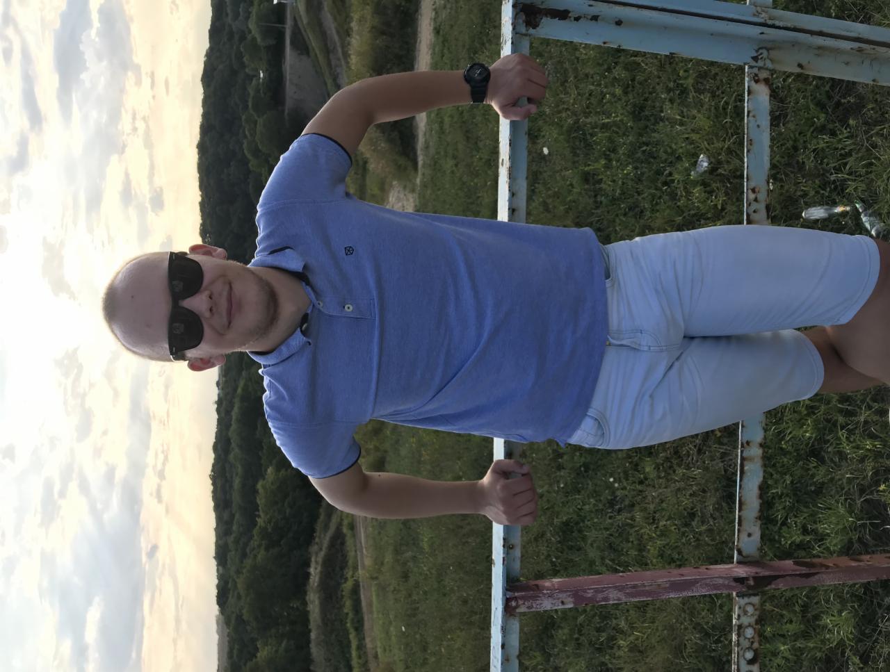 Mężczyzna w okularach, opierający się o metalową barierkę na tle zieleni i nieba. Ubrany w niebieską koszulkę polo i jasne szorty. Widok z bliska, z lekkiego ukosa.