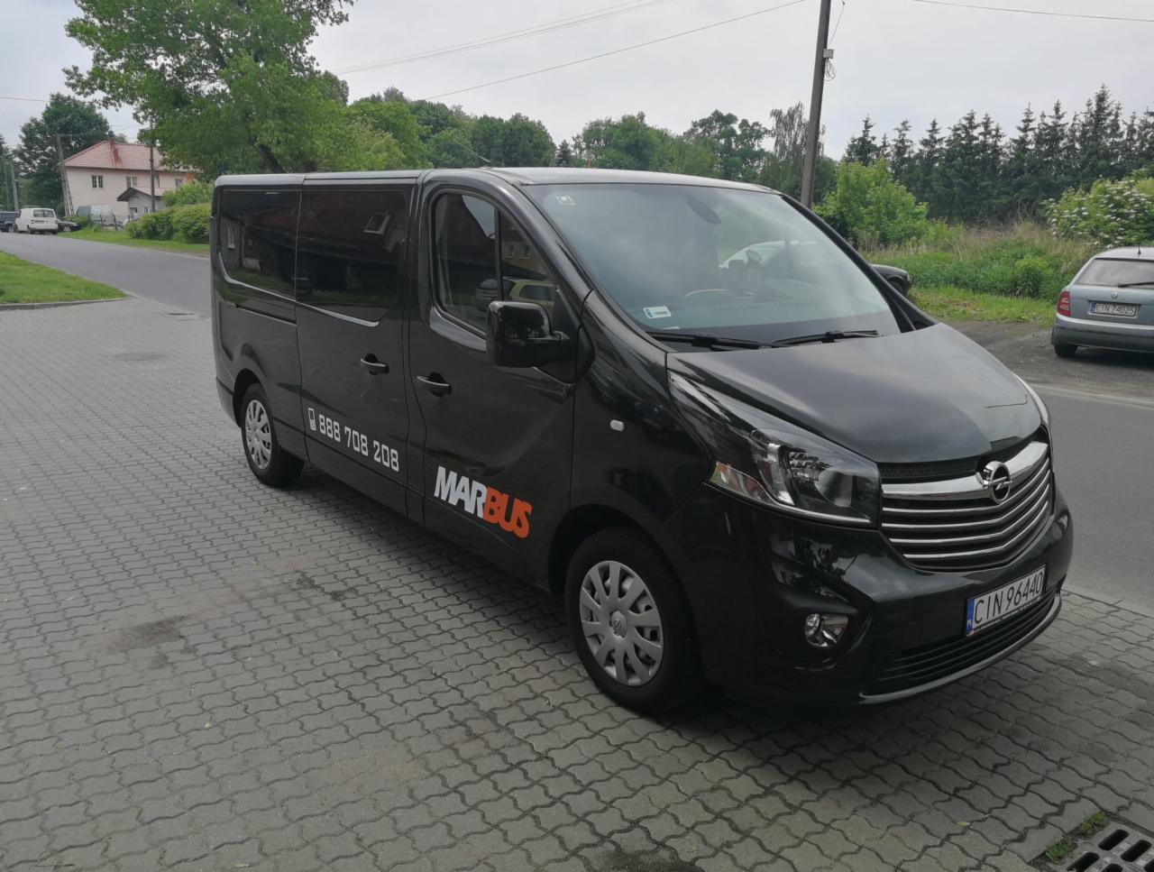 Czarny bus Opel Vivaro z logo Marbus i numerem telefonu zaparkowany na kostce brukowej przed budynkiem.