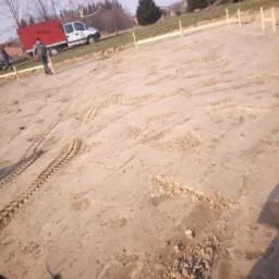 Przygotowany teren pod budowę z wytyczonymi drewnianymi słupkami, widoczne ślady opon, w tle samochody dostawcze i drzewa.