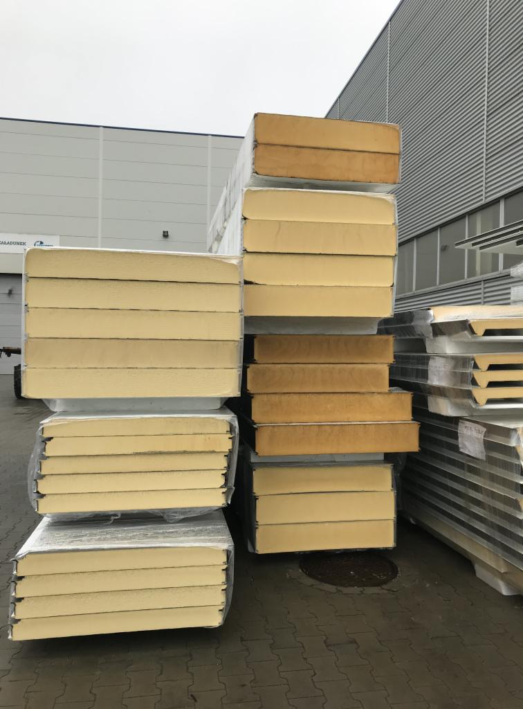 Stosy paneli warstwowych w folii ochronnej, ułożone na zewnątrz magazynu z szarą elewacją z blachy trapezowej.