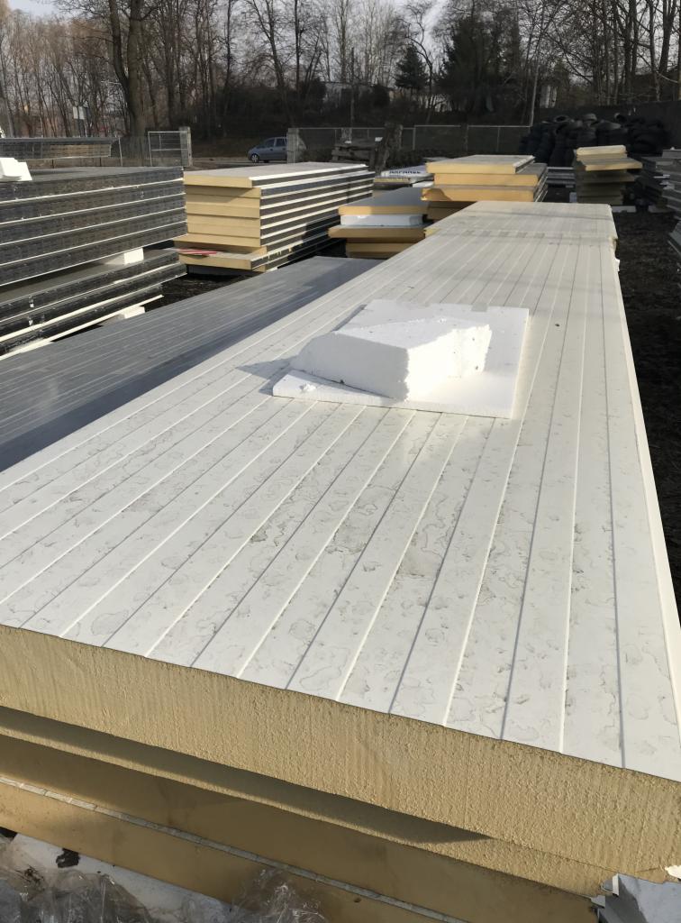 Stos płyt warstwowych z widocznym rdzeniem izolacyjnym i białą, rowkowaną powierzchnią zewnętrzną, na wierzchu leży biały blok styropianu, w tle składowane inne płyty i drzewa.