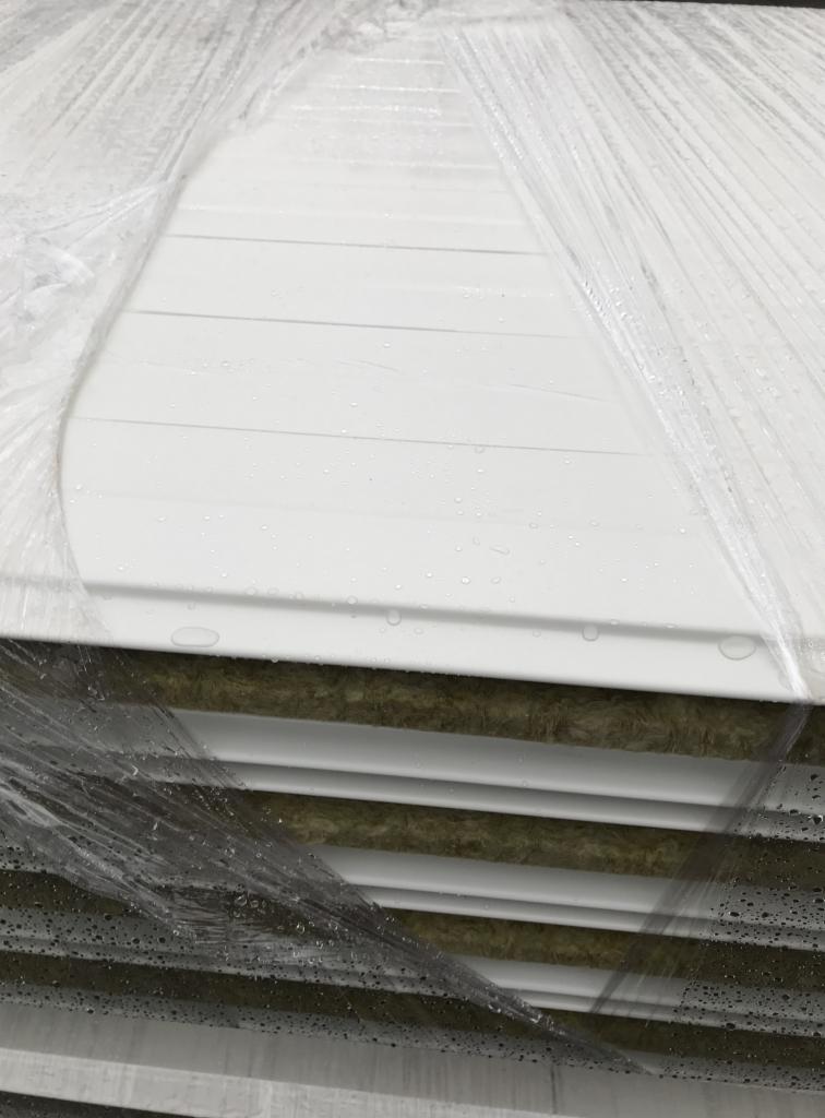 Stos białych płyt warstwowych z widocznym wypełnieniem izolacyjnym, zabezpieczonych folią ochronną z kroplami wody.