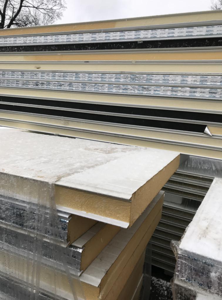 Stos płyt warstwowych z widocznym rdzeniem izolacyjnym, zabezpieczonych folią, składowanych na zewnątrz pod pochmurnym niebem.