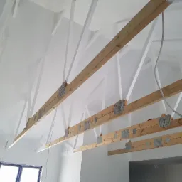 Poddasze w trakcie prac: drewniane belki stropowe z metalowymi łącznikami, częściowo pomalowane na biało, widoczne przewody elektryczne i drabina oparta o ścianę.