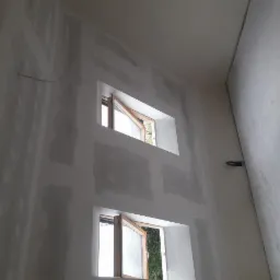 Wnętrze w trakcie remontu: ściany z zamontowanymi oknami, częściowo pokryte masą szpachlową, przygotowane do dalszych prac wykończeniowych.