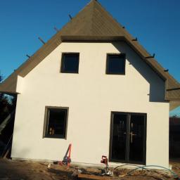 Marcin Piekarski - Surowy stan domu z białymi ścianami i ciemnymi oknami, widoczne narzędzia budowlane i drewno na placu budowy, dach w trakcie wykańczania z widocznymi krokwiami.