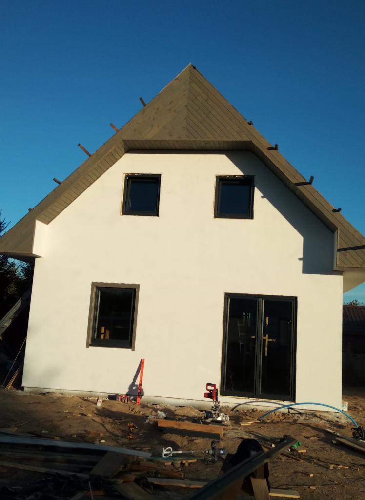 Surowy stan domu z białymi ścianami i ciemnymi oknami, widoczne narzędzia budowlane i drewno na placu budowy, dach w trakcie wykańczania z widocznymi krokwiami.