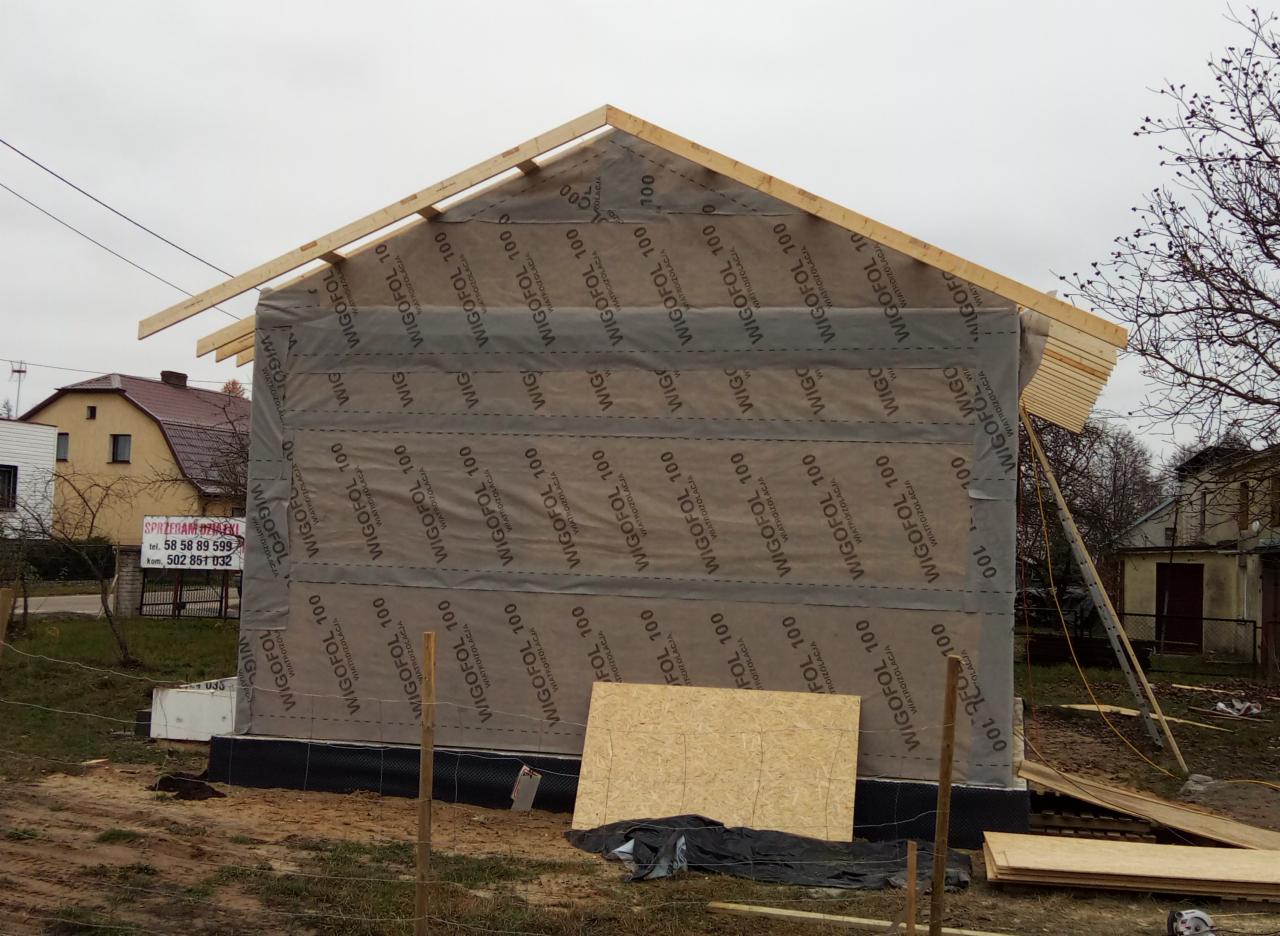 Wznoszony dom w trakcie budowy, zabezpieczony folią wiatroizolacyjną z widocznymi drewnianymi elementami konstrukcyjnymi dachu i ścian, na pierwszym planie płyta OSB.