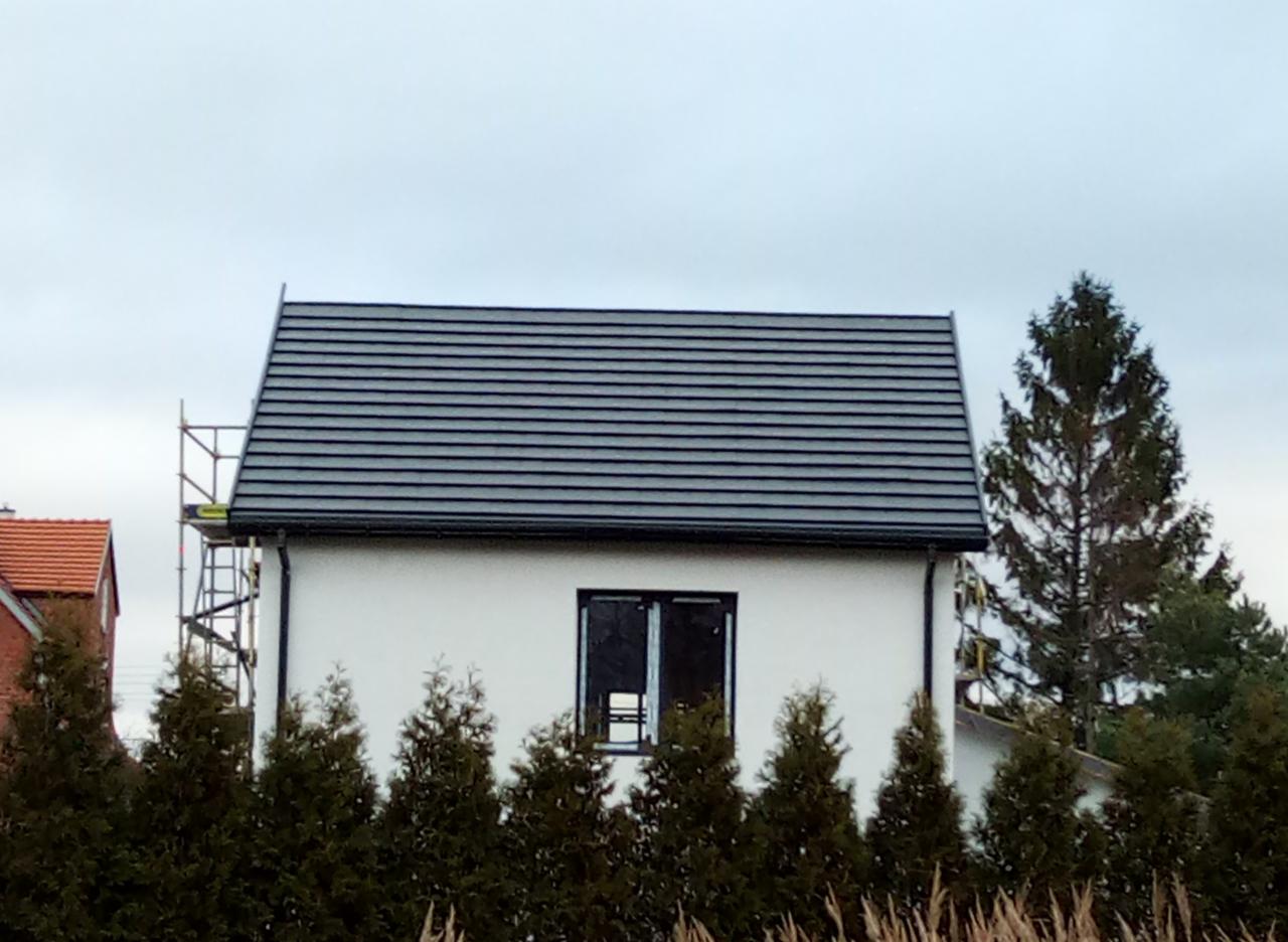 Biały dom z ciemną dachówką, widoczne rusztowanie z boku, otoczony iglakami i trawami.