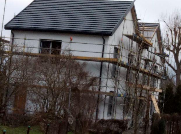 Dwupiętrowy dom w trakcie ocieplania elewacji, z szarym dachem i rusztowaniami, widoczny przez gałęzie drzew.