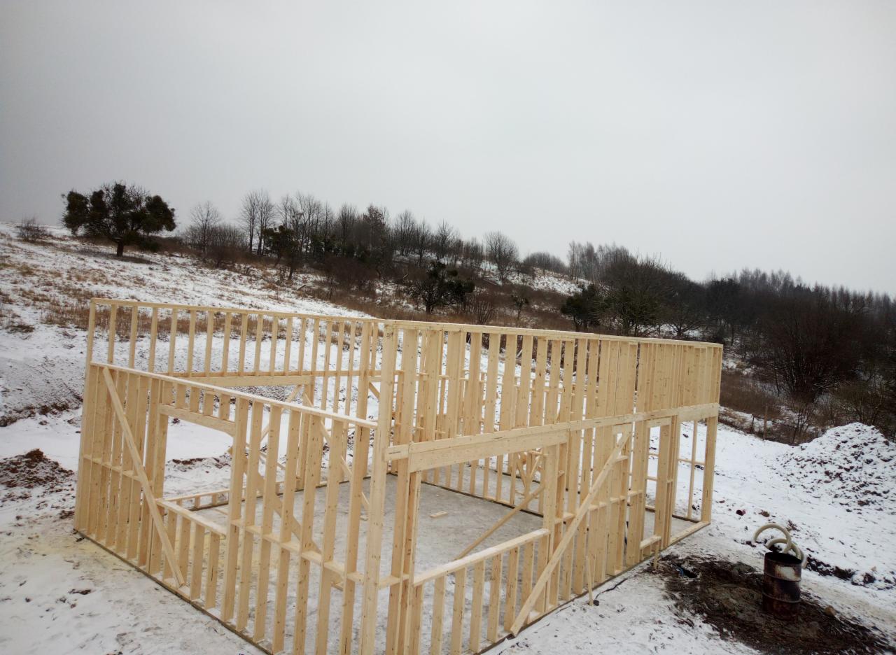 Konstrukcja szkieletowa domu w trakcie budowy, widok na wzgórze z drzewami w zimowej scenerii, z widocznym starym, zardzewiałym bakiem na pierwszym planie.