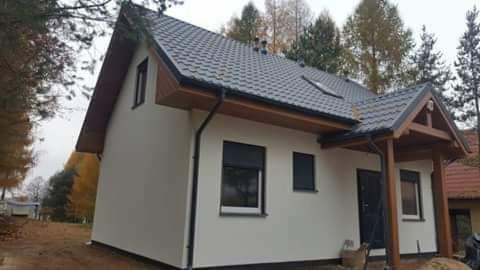 Gotowy dom jednorodzinny z białymi ścianami, grafitowym dachem z lukarną i drewnianą wiatą nad wejściem, otoczony drzewami.