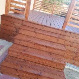 Nowy, drewniany taras z balustradą i schodami, wykończony brązowym lakierem, widok z dołu na tle zieleni.