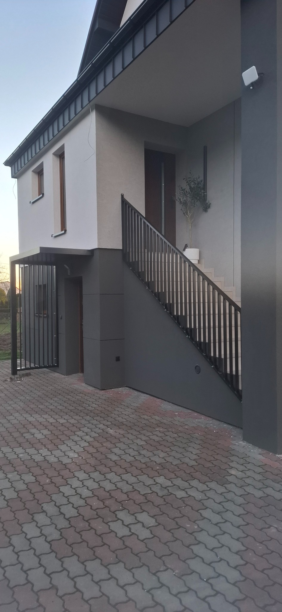 Nowoczesny dom z szarą metalową balustradą schodową i bramką wejściową. Minimalistyczny design, kostka brukowa na podjeździe, drzewko w donicy przy schodach.