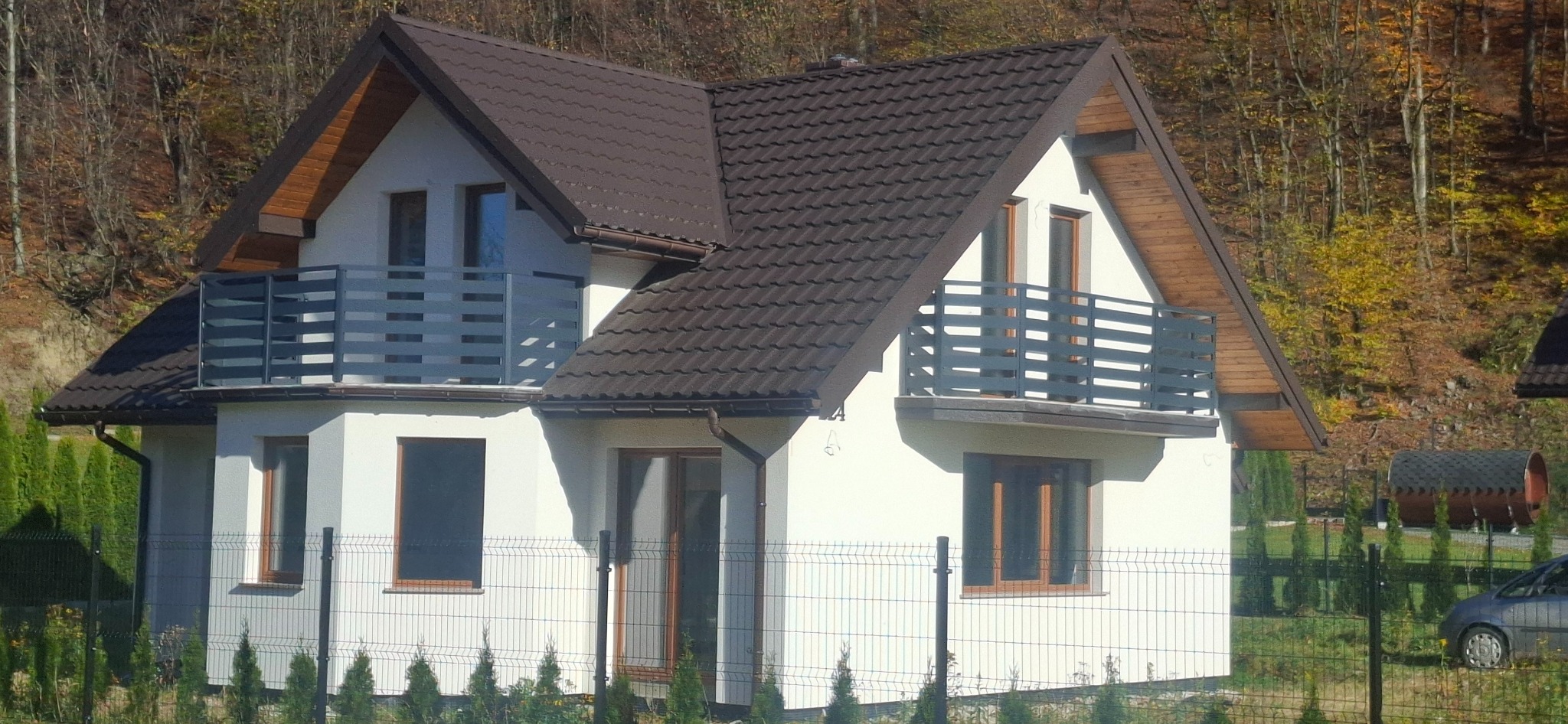 Nowoczesny dom z balkonami zabezpieczonymi szarymi, metalowymi balustradami o poziomym układzie szczebli, na tle jesiennego lasu.