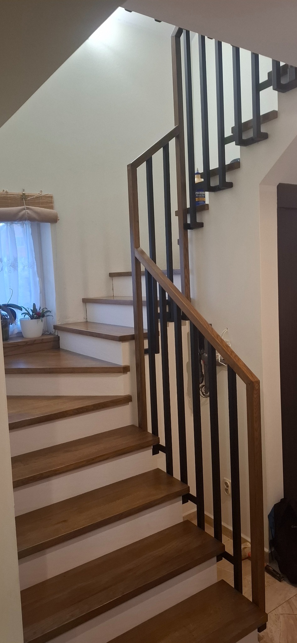 Drewniane schody z czarną, metalową balustradą w nowoczesnym domu. Widoczne stopnie z jasnym obramowaniem i rośliny w doniczce przy oknie.