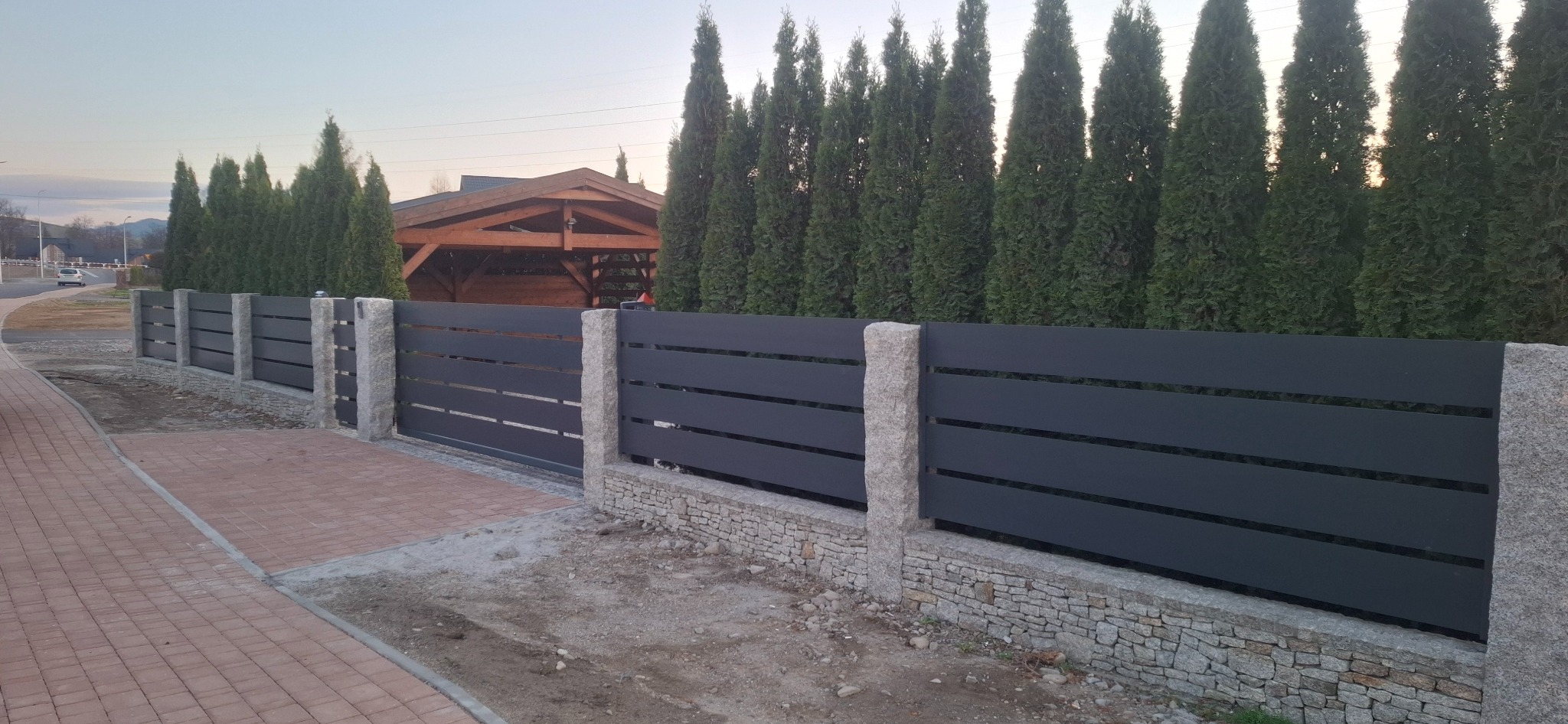 Nowoczesne ogrodzenie metalowe z poziomymi panelami w kolorze antracytowym i kamiennymi słupkami, na tle zielonych tuj i drewnianej altany.