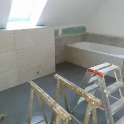 Remont łazienki na poddaszu: zabudowa WC z płytkami o falistej strukturze, obudowa wanny z szarymi płytkami, widoczne przygotowane instalacje i drabiny malarskie.