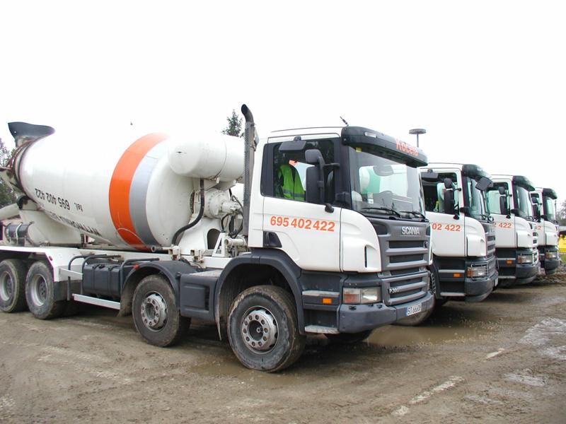 Szereg białych betonomieszarek Scania P380 z pomarańczowo-szarymi pasami na zbiornikach, zaparkowanych na błotnistym placu budowy.