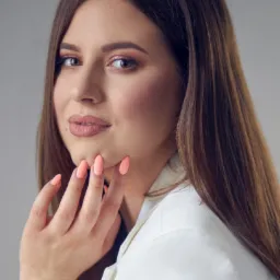 Portret kobiety z lekkim uśmiechem, z delikatnym upiększeniem oczu w odcieniach różu i brzoskwini, oraz z pomadką w kolorze nude, dłoń z pomalowanymi na brzoskwiniowo paznokciami dotyka policzka...