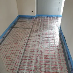 Rozłożona instalacja ogrzewania podłogowego z czerwonymi rurkami na siatce, zabezpieczona niebieską folią przy ścianach, widok z góry, surowy stan wykończenia.