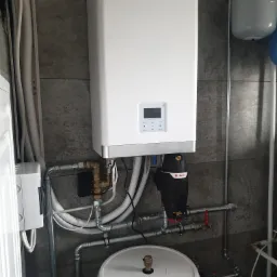 Biały piec gazowy marki Sevra zamontowany na szarej, teksturowanej ścianie, z widocznymi rurami, zaworami i filtrem Flamco.