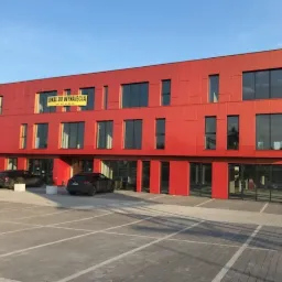 Nowoczesny, dwupiętrowy budynek biurowy z czerwonymi panelami elewacyjnymi i dużymi oknami, z żółtą tablicą 'LOKAL DO WYNAJĘCIA' na drugim piętrze, zaparkowane samochody na brukowanym parkingu...