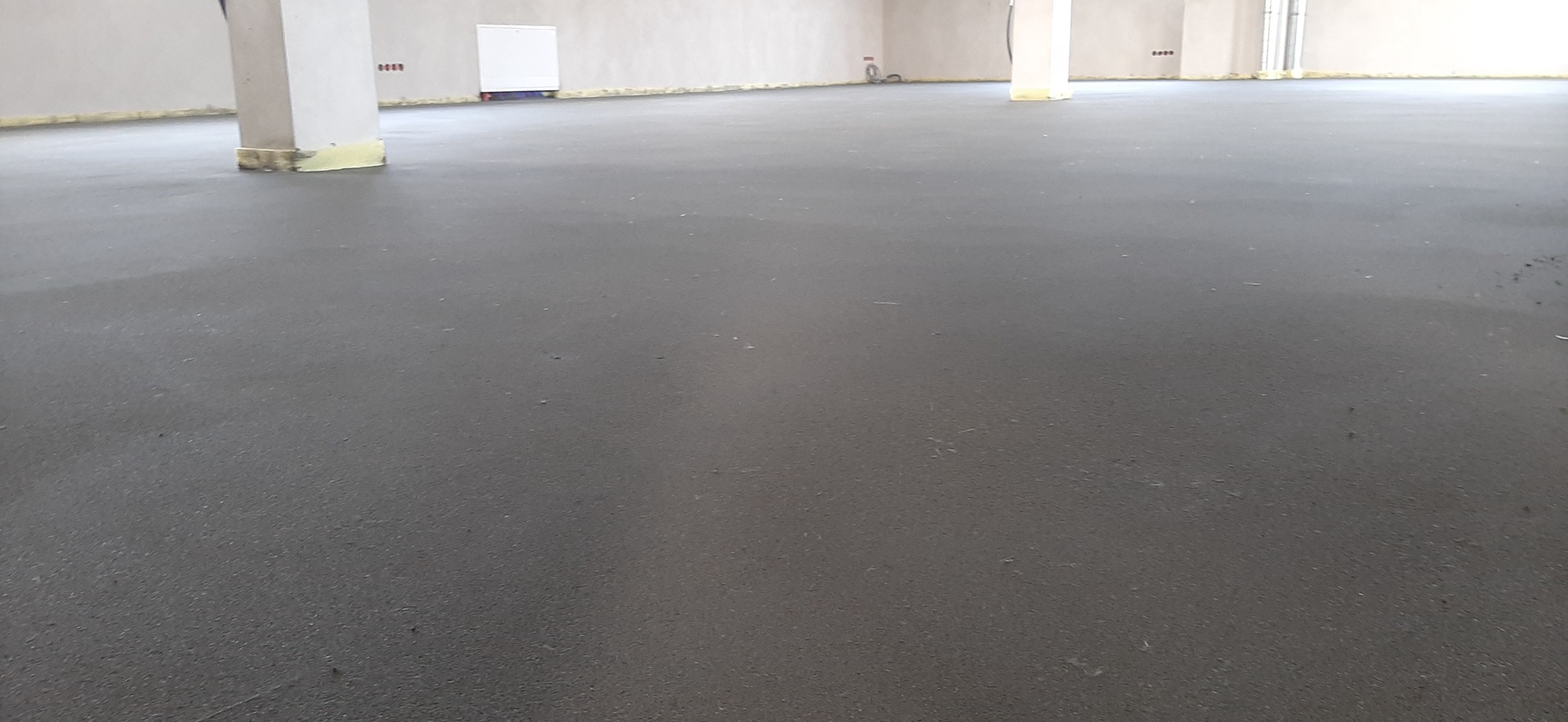 Świeżo wylana, gładka, szara posadzka w dużym pomieszczeniu z widocznymi słupami konstrukcyjnymi i przygotowanymi ścianami do dalszych prac wykończeniowych.