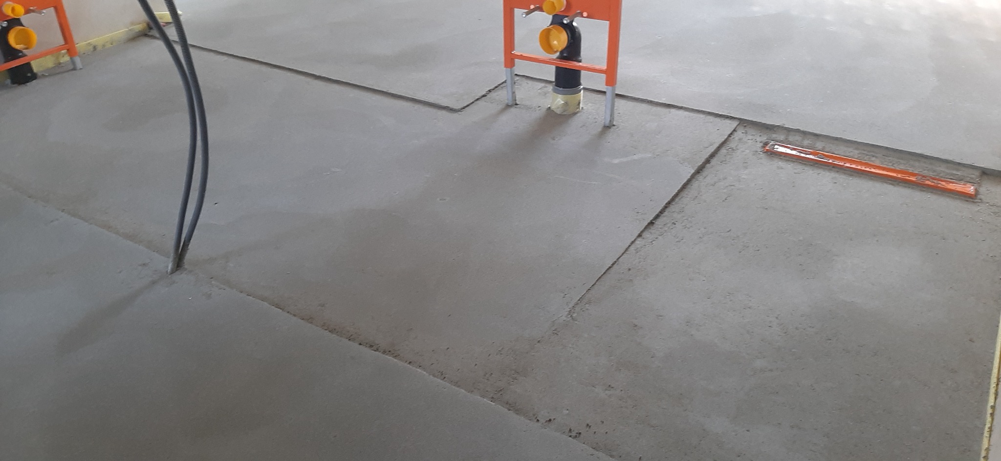 Świeżo wylana posadzka jastrychowa z widocznymi elementami instalacji sanitarnych i elektrycznych, pomarańczowa listwa dylatacyjna w tle.