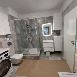 Sławomir Pożarowski - Wykończona łazienka z kabiną prysznicową, umywalką z szafką i lustrem z podświetleniem LED. Na podłodze szare płytki imitujące kamień. Widoczna pralka i białe szafki.