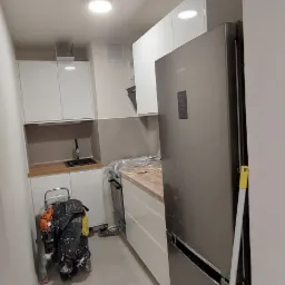 Wąska kuchnia w trakcie remontu: białe szafki, szary sprzęt AGD, drabina malarska z ubraniami roboczymi, podłoga wyłożona jasnymi płytkami.