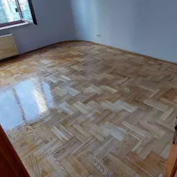 Parkiet ułożony w jodełkę, widoczny przez otwarte drewniane drzwi z szybą o wzorzystej fakturze, w tle okno z widokiem na wysokie budynki.