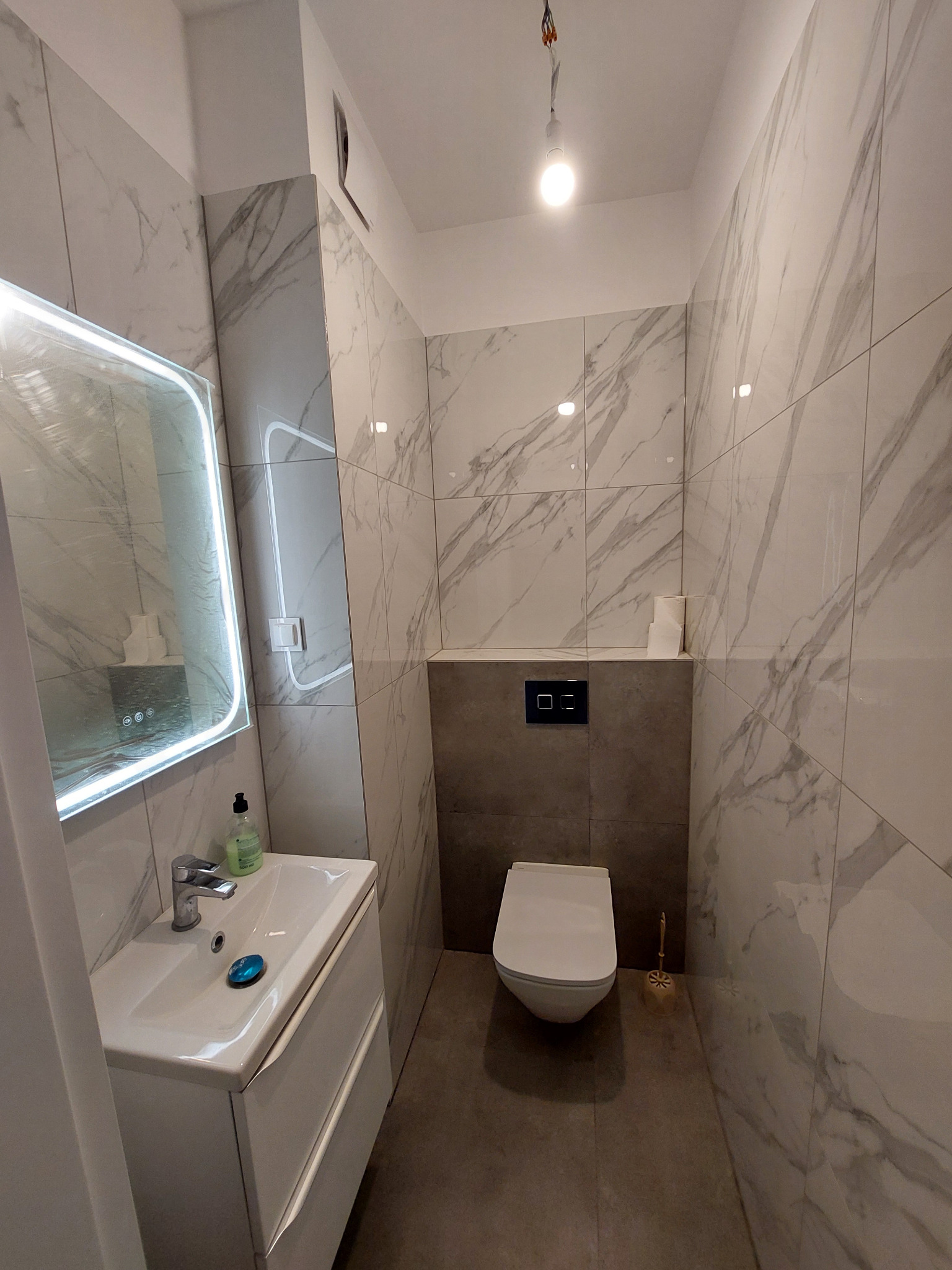 Nowoczesna łazienka z marmurowymi płytkami, podświetlanym lustrem nad białą szafką z umywalką i wiszącą toaletą. Minimalistyczny design, szare akcenty.