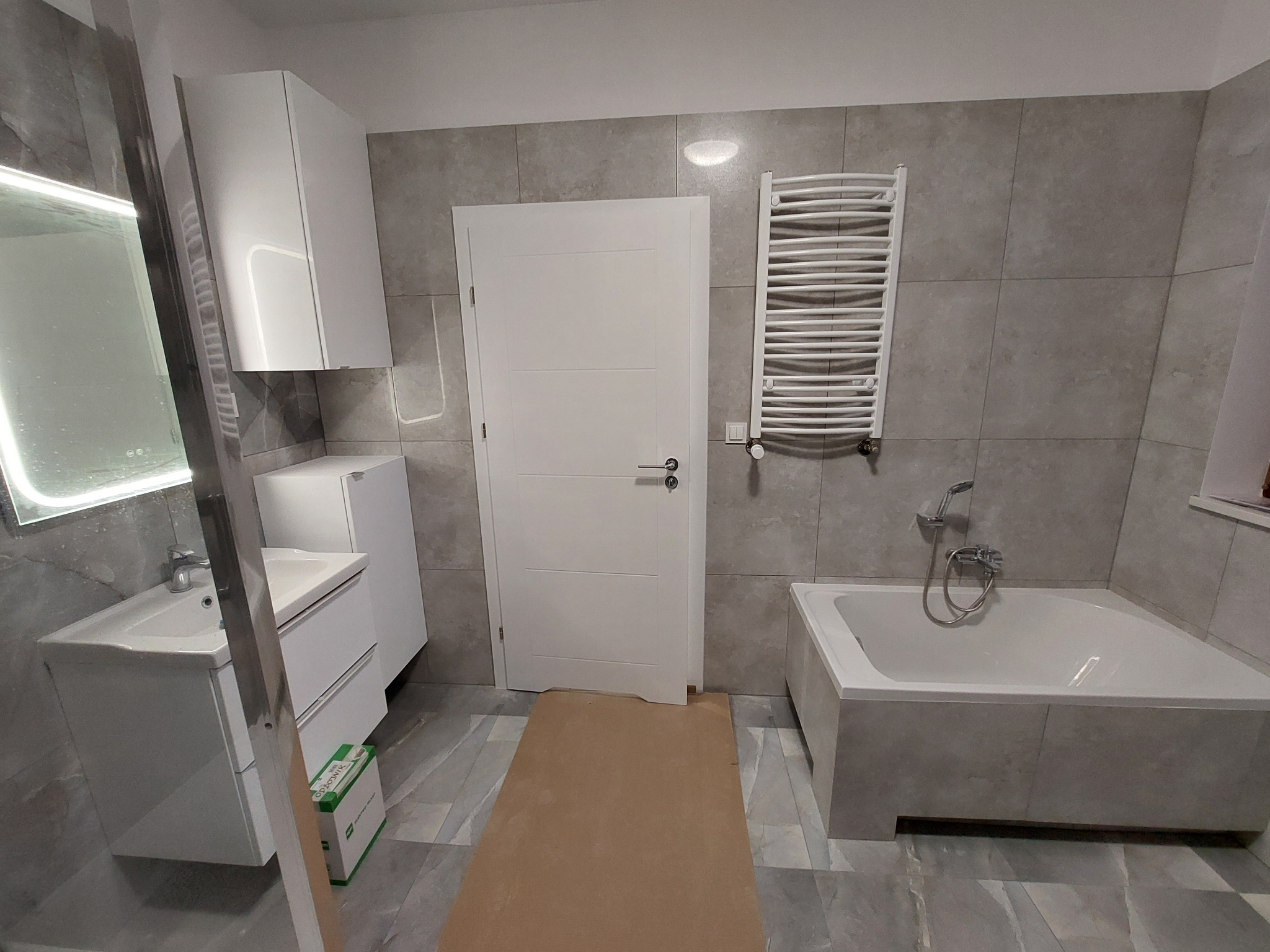 Jasna łazienka po remoncie: szare płytki, biała armatura (umywalka z szafką, wanna), grzejnik drabinkowy. Na podłodze zabezpieczenie. Minimalistyczny design.