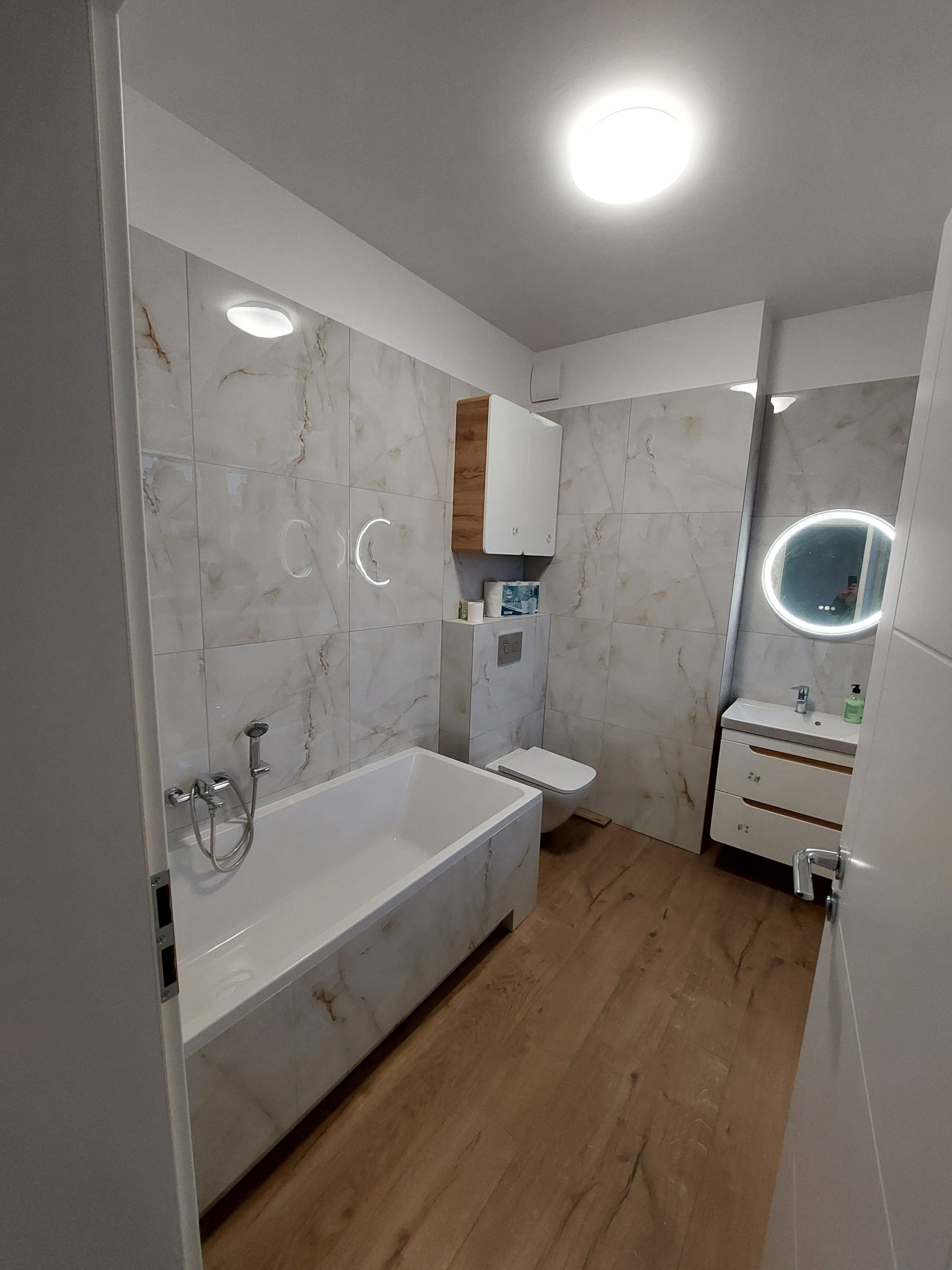 Jasna łazienka po remoncie z wanną, toaletą, umywalką z szafką i okrągłym lustrem z podświetleniem LED. Płytki imitujące marmur na ścianach i drewnopodobna podłoga.