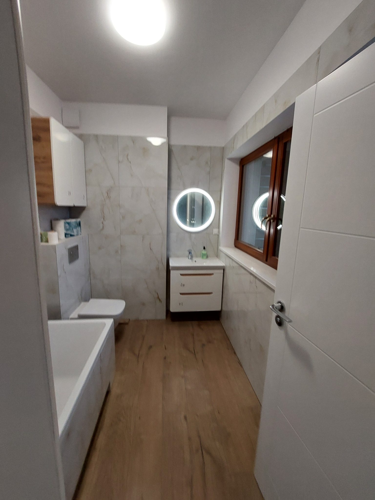 Jasna łazienka po remoncie: drewnopodobne płytki, marmurowe ściany, okrągłe lustro z podświetleniem, biała armatura i szafki. Widok z perspektywy wejścia.