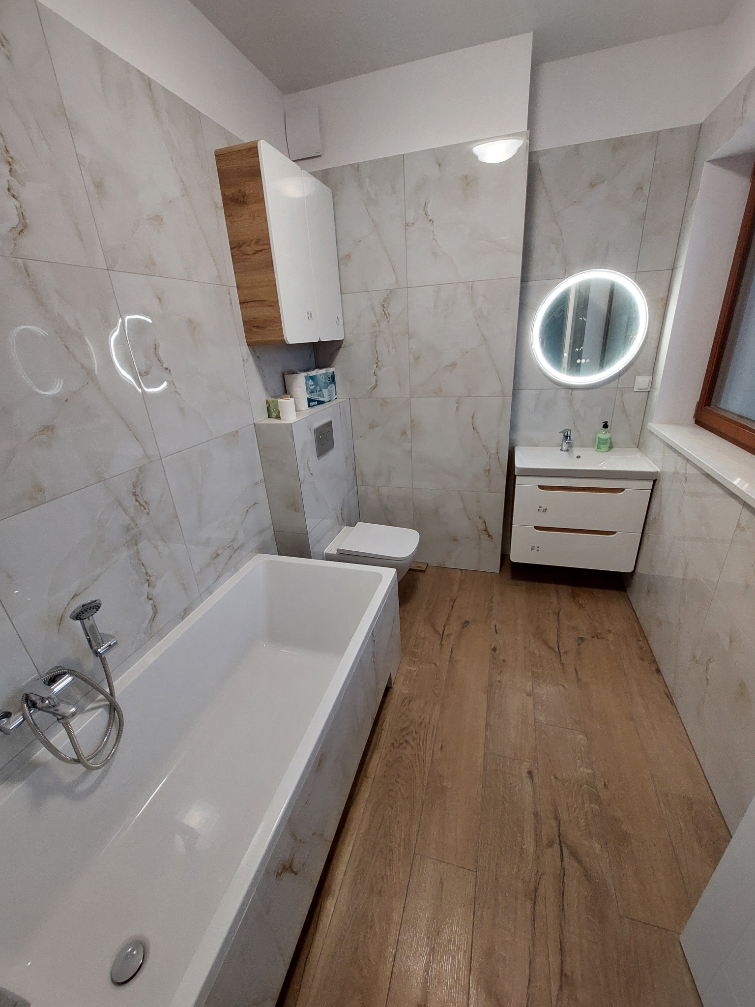 Wykończona łazienka z wanną, toaletą, umywalką z szafką i okrągłym lustrem z podświetleniem. Płytki imitujące marmur na ścianach, drewnopodobna podłoga.