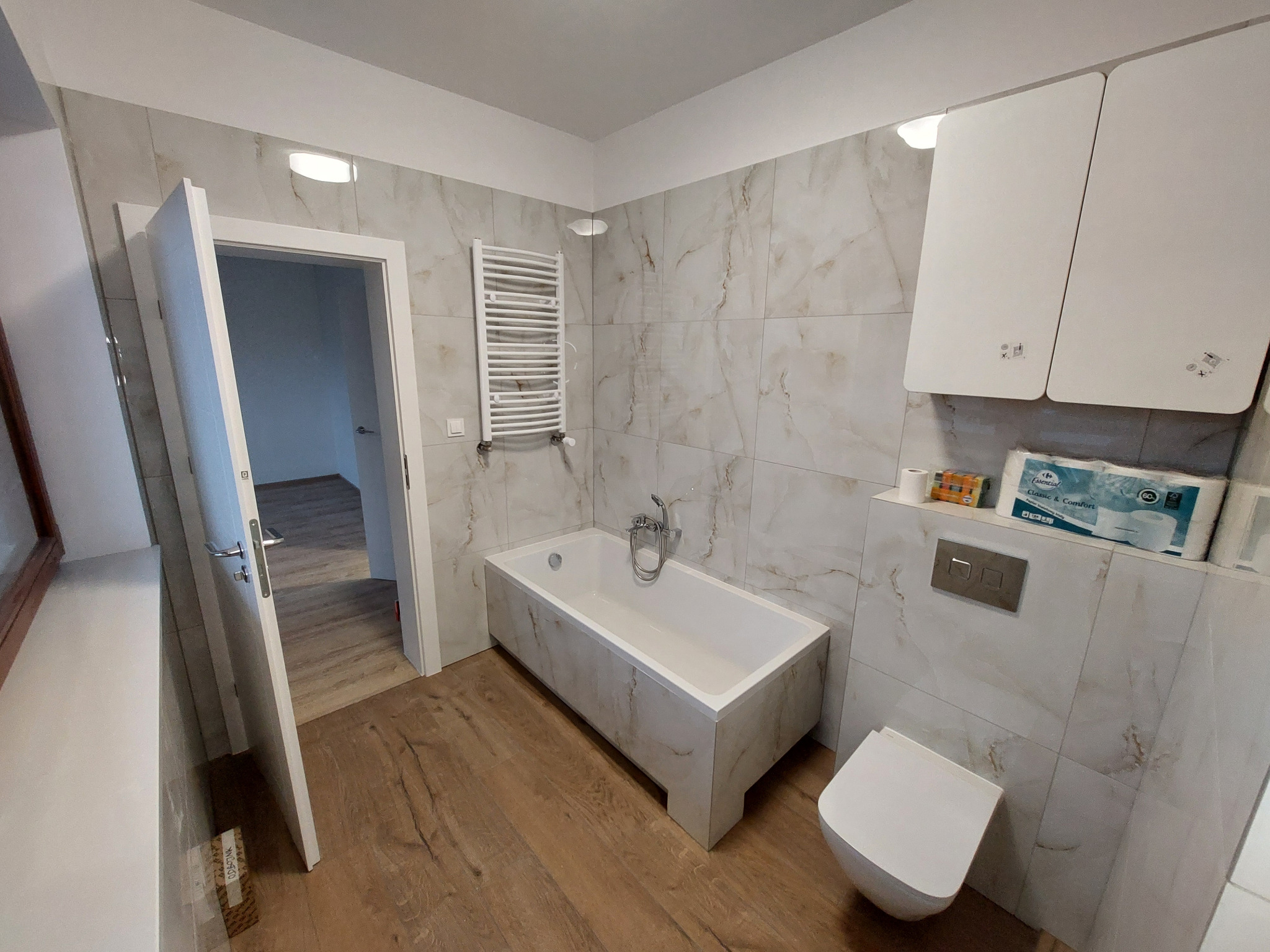 Jasna łazienka po remoncie: wanna obudowana płytkami imitującymi marmur, drewnopodobna podłoga, biała toaleta i szafki. Widoczny grzejnik drabinkowy.