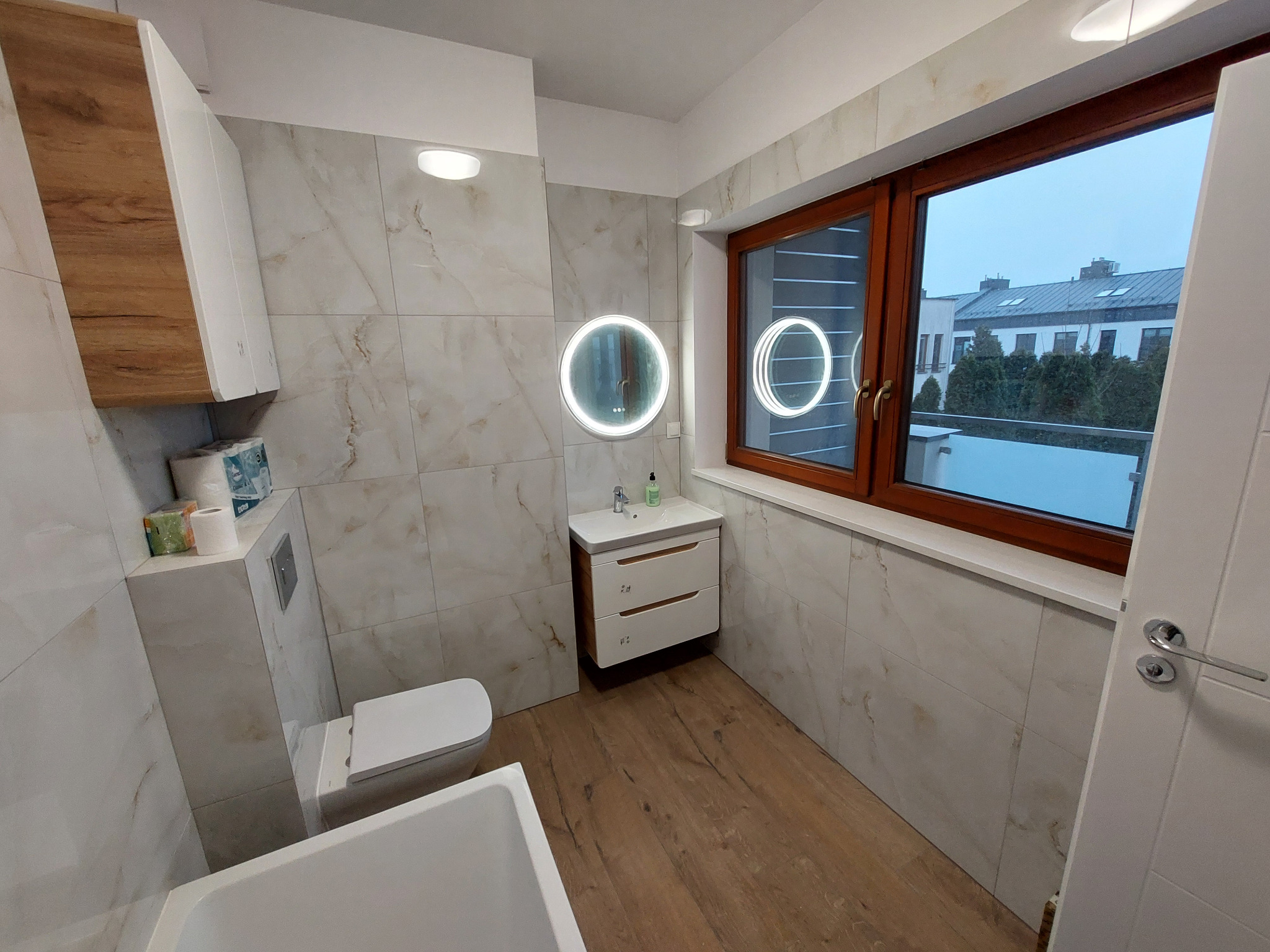 Jasna łazienka z drewnianą podłogą i oknem. Umywalka z szafką, toaleta w zabudowie, lustro z podświetleniem. Płytki imitujące marmur na ścianach.