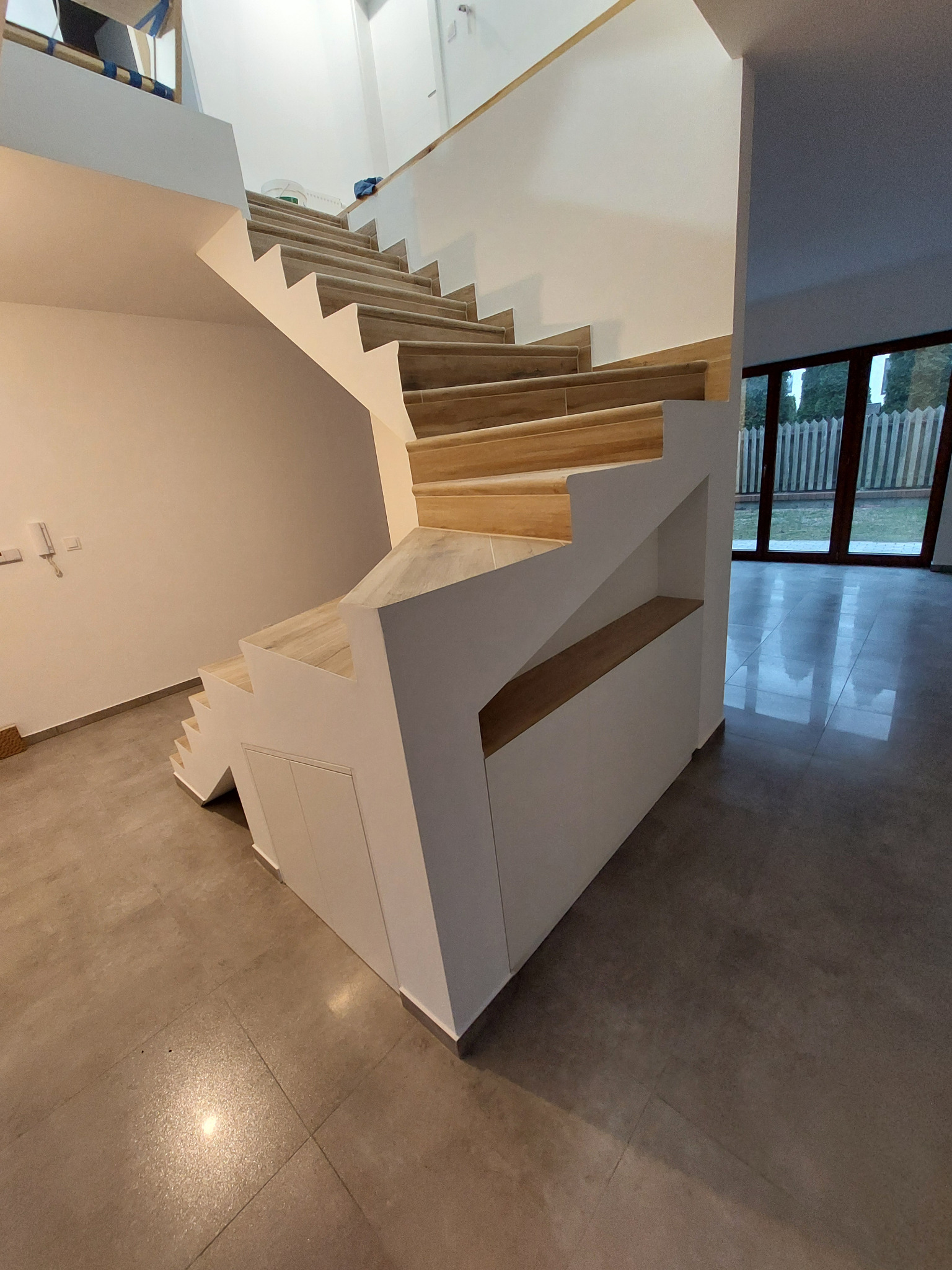 Nowoczesne schody z drewnianymi stopniami i białymi bokami, z wbudowanymi szafkami i półkami, na tle jasnego wnętrza z dużymi oknami.