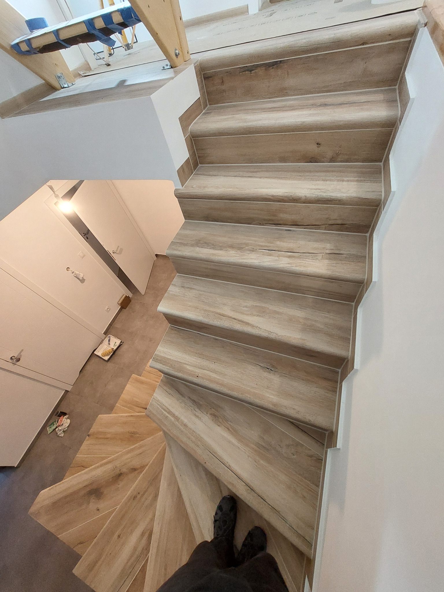 Wykończone schody z płytek imitujących drewno, widok z góry, z fragmentem osoby w kadrze. Białe ściany i drzwi w tle. Nietypowy kąt ujęcia.