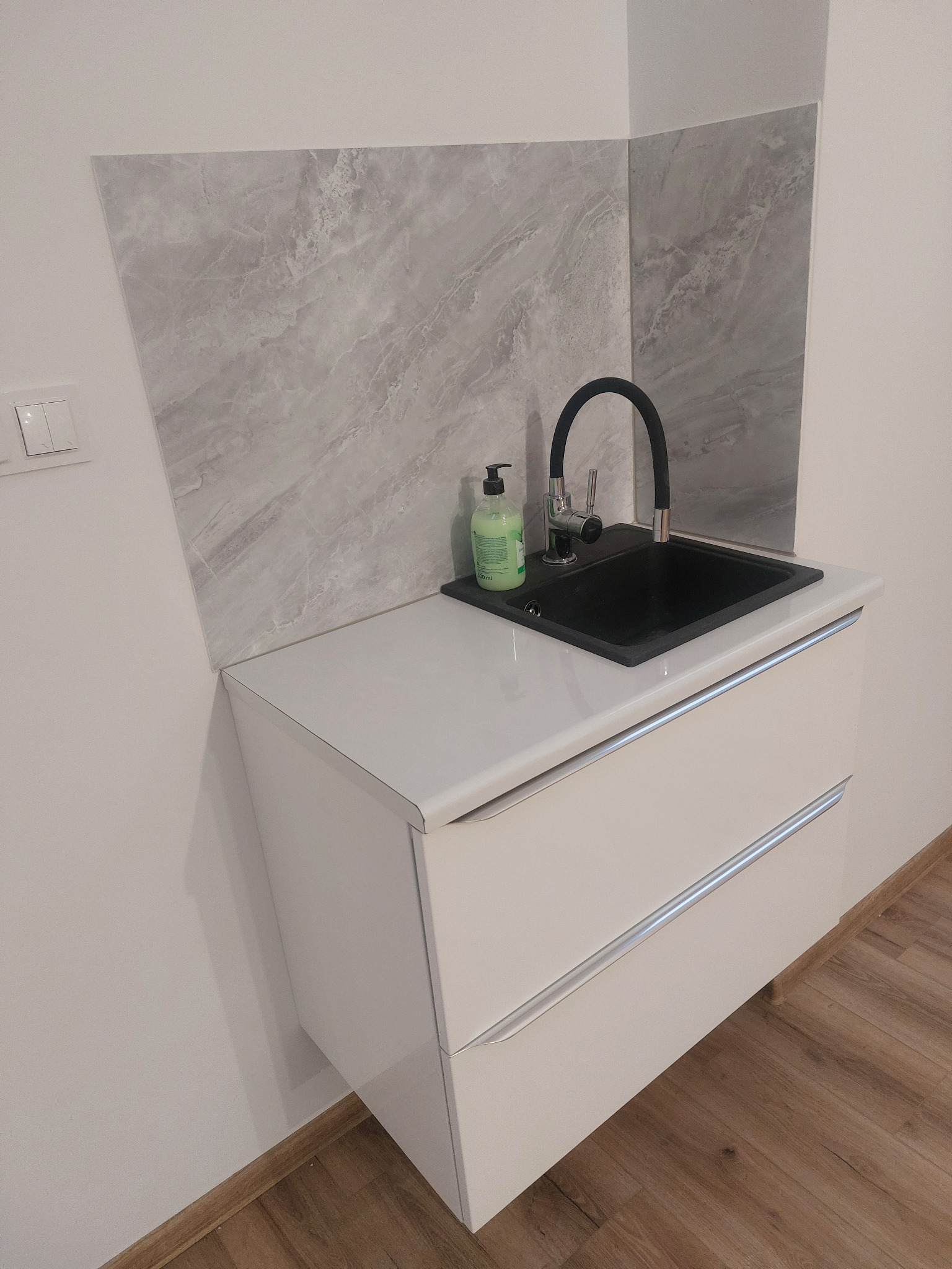 Nowoczesna łazienka: czarna umywalka z baterią na białej szafce z szufladami, marmurowe płytki na ścianie. Minimalistyczny design, butelka mydła w tle.