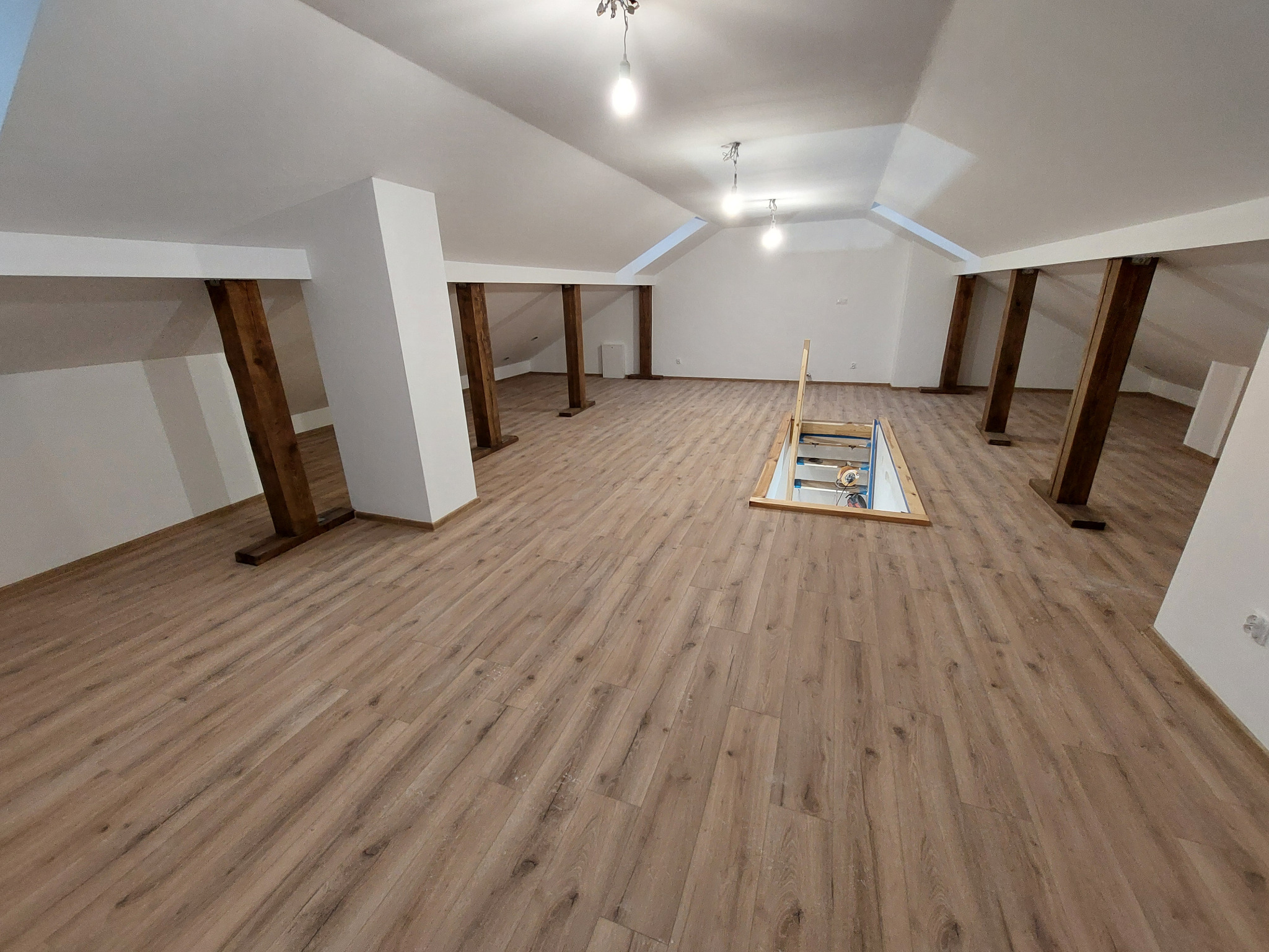 Jasne poddasze z drewnianymi belkami i panelami podłogowymi. Widoczne schody strychowe i oświetlenie sufitowe. Przestronne wnętrze po remoncie.