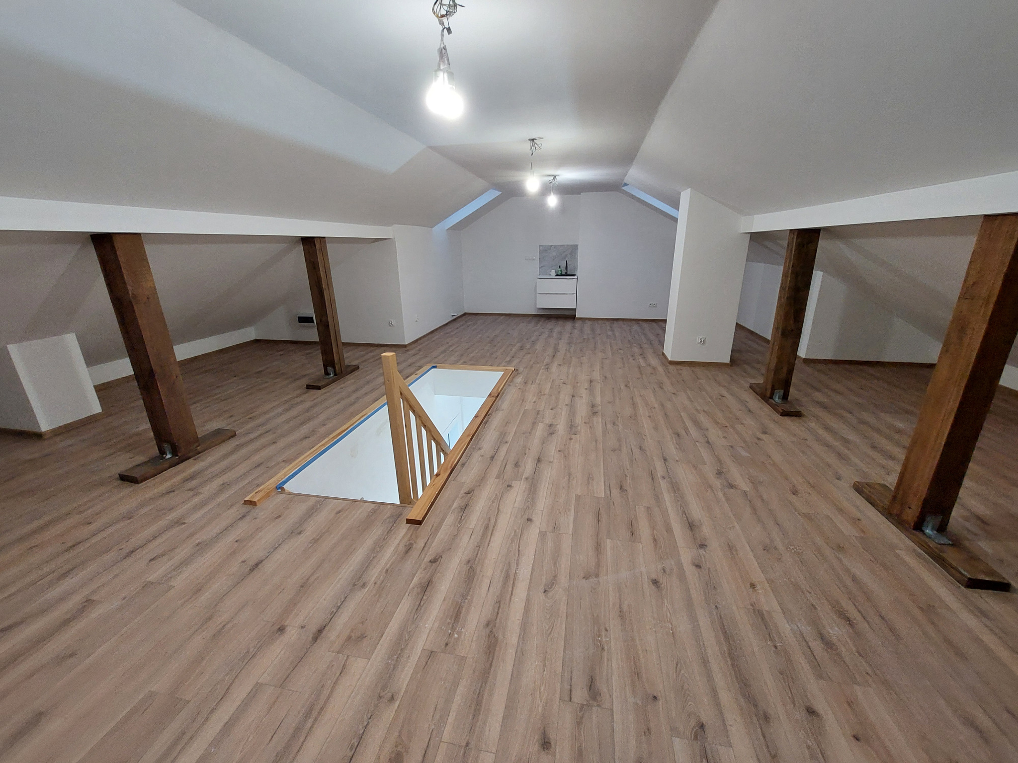 Jasne poddasze z drewnianą podłogą i schodami. Widoczne drewniane belki konstrukcyjne i białe ściany. W tle mała łazienka z białą szafką i umywalką.