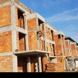 Surowy stan budynku wielorodzinnego z czerwonej cegły i betonu, widoczne drewniane elementy konstrukcyjne dachu, w tle błękitne niebo i zabudowa jednorodzinna.