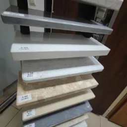 Ekspozycja próbek różnych kolorów i wzorów parapetów, ułożonych jeden na drugim, z widocznymi etykietami z nazwami: Olympo, New Marfil, Baltic Grey, Perlato Royal, Misty White, Polare.