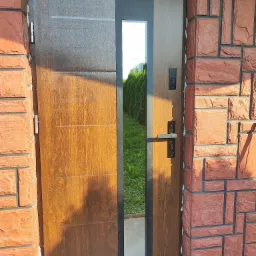 Brązowe drzwi wejściowe z szybą, zamontowane w czerwonej ceglanej elewacji, widoczne ślady pianki montażowej.