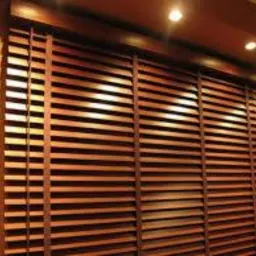 Zasłonięte okno z drewnianymi żaluzjami w ciepłym odcieniu brązu, widoczne poziome lamele i sznurki sterujące, oświetlone punktowymi lampami sufitowymi, na ścianie w kolorze beżowym.